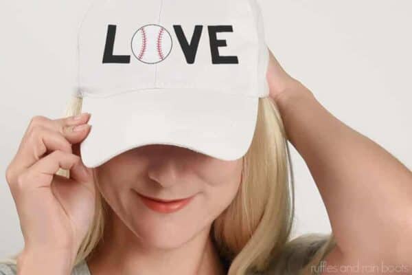 Horizontal image of blonde, smiling woman in white baseball hat with LOVE baseball SVG done in black, red, and white vinyl.