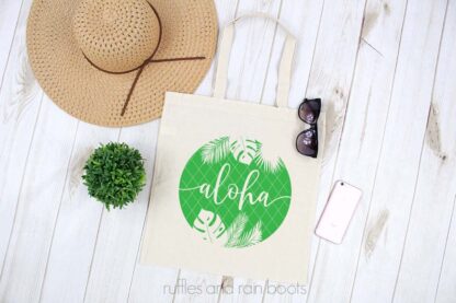 Horizontal image showing a canvas tote bag with green vinyl of aloha summer SVG with sunglasses, a phone, and a sun hat on white wood background.