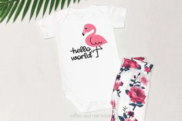 Horizontal image of linen background with fern, floral pants, and white bodysuit with pink and black flamingo SVG in cute Kawaii style which reads hello world.