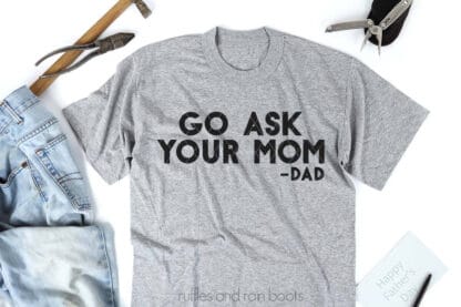 Horizontal image of a light gray tee on white background with black vinyl which reads go ask your mom -dad.