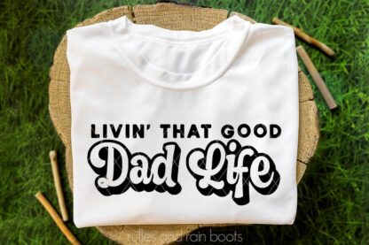 Horizontal image close up of a white t shirt folded on a stump sitting in grass with black vinyl which reads livin that good dad life.