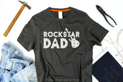 Horizontal image of dark gray shirt with gray vinyl rockstar dad svg with guitar on white background with jeans and tools.