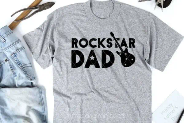 Horizontal image of light gray t-shirt with black rockstar dad svg in vinyl on white background with jeans and tools.