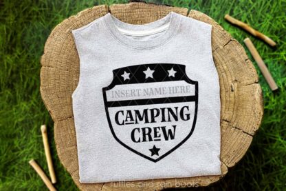 Horizontal image of a folded white t-shirt with a badge camping crew SVG in black vinyl on stump with grass background.