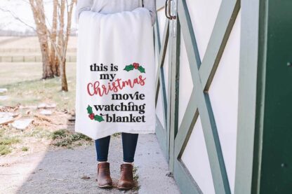 Horizontal image of a woman next to a barn holding a white blanket which reads This is my Christmas movie watching blanket in heat transfer vinyl.