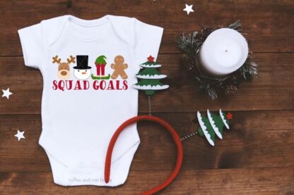 Horizontal image of Christmas squad goals SVG on white baby bodysuit on dark wood background.