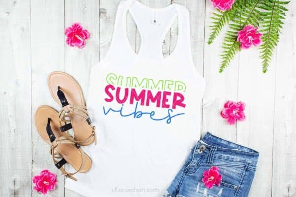 Horizontal image of shorts, sandals, and white tank top with neon stacked summer vibes SVG in heat transfer vinyl on tropical wood background.