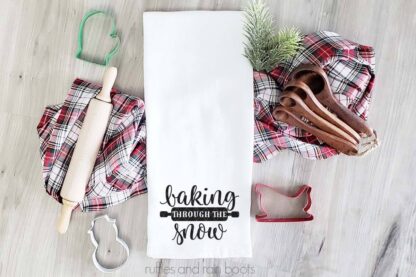 Horizontal image of a white kitchen towel with heat transfer vinyl saying baking through the snow with a rolling pin on a holiday backgorund.
