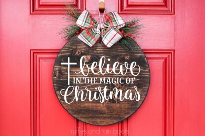 Horizontal image of a believe in the magic of Christmas svg in white vinyl on dark wood background against a bright red door.