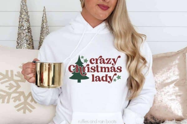 Horizontal image of a woman in front of holiday pillows holding a gold cup wearing a white sweatshirt with crazy Christmas lady in vinyl.