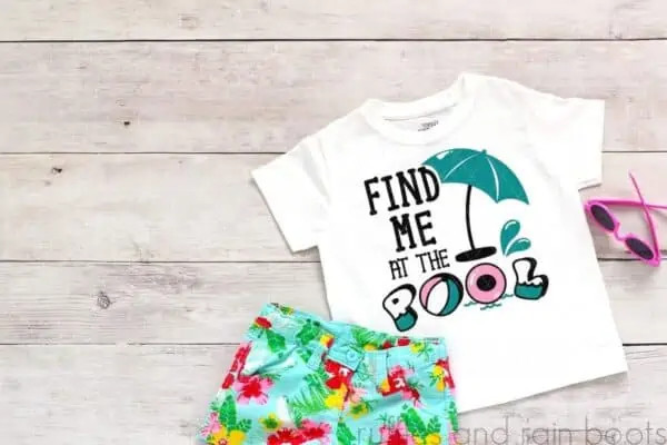 Horizontal image of a white t-shirt with teal, pink, and black vinyl which reads find me at the pool on light wood background with summer accents.