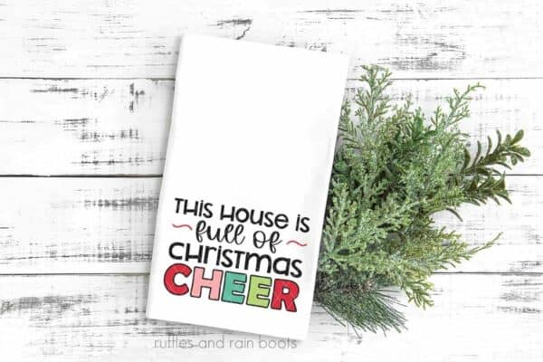 Horizontal image of white kitchen towel on wood background with This House is Full of Christmas Cheer SVG in colorful heat transfer vinyl.