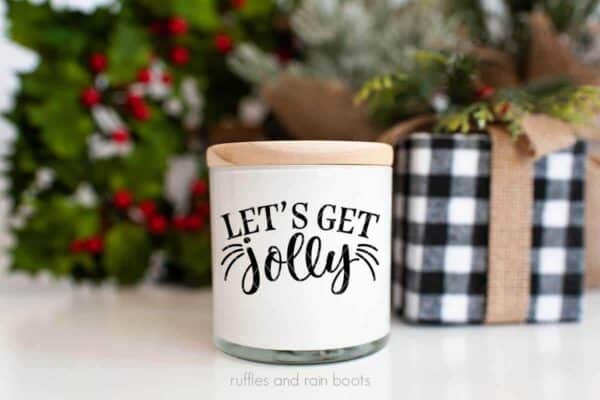 Horizontal image of a white candle with wood lid and black vinyl which reads let's get jolly in front of Christmas backgound.
