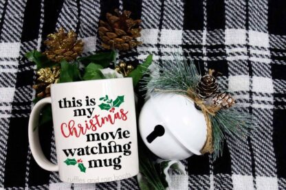 Horizontal image of a Buffalo check fabric with a white bell and white coffee cup with vinyl which reads this is my Christmas movie watching mug.