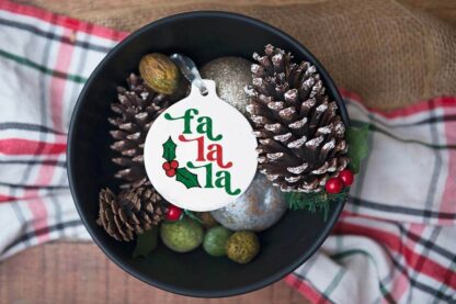 White porcelain ornament with fa la la SVG with holly and berry accent in bowl with filler on holiday table.