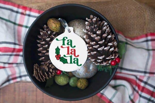 White porcelain ornament with fa la la SVG with holly and berry accent in bowl with filler on holiday table.