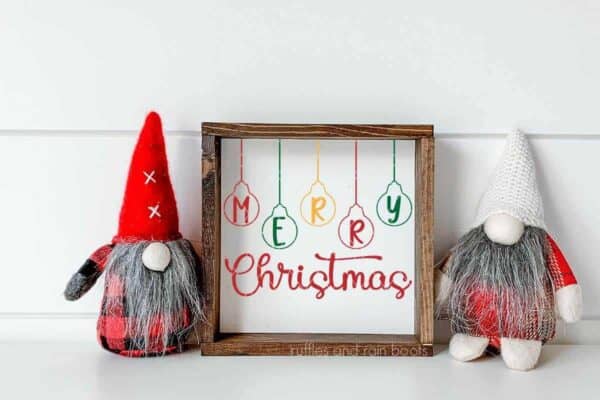 Horizontal image of two gnomes standing either side of a square frame with free Merry Christmas SVG with ornaments.