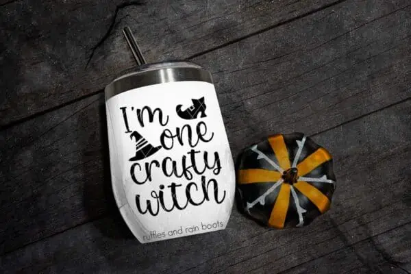 Horizontal image of a white tumbler with I'm one crafty witch SVG with witch shoe and witch hat in black vinyl next to a painted pumpkin on black wood.
