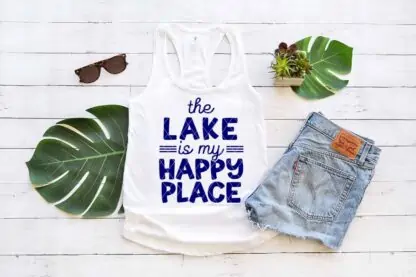 Horizontal image of white wood, monstera leaves, and white tank which reads the lake is my happy place in blue heat transfer vinyl made with Cricut.