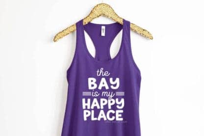 Horizontal image of a gold glitter hanger holding a purple tank top with white vinyl letters which read the bay is my happy place.