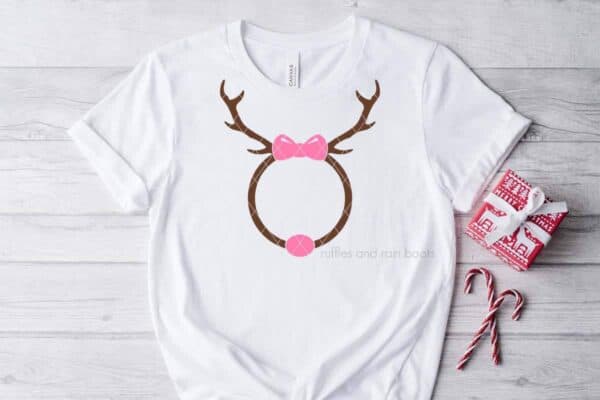 Horizontal image of a white t-shirt with a reindeer monogram SVG with a pink bow and nose on light wood background with holiday accents.