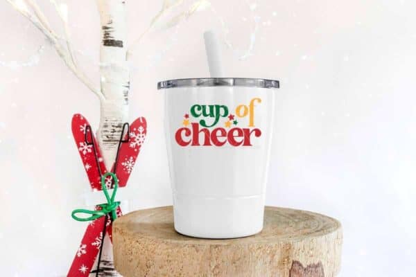 White toddler tumbler which reads cup of cheer with stars on a wood plank next to a pair of mini skis.