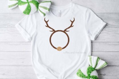 Horizontal image of a white t-shirt with a reindeer monogram SVG with a light brown nose on light wood background with holiday accents.
