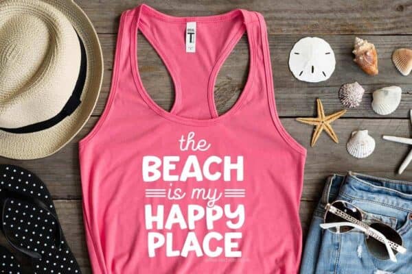 Horizontal image of wood background with coastal elements and a pink tank top which reads the beach is my happy place in white vinyl.