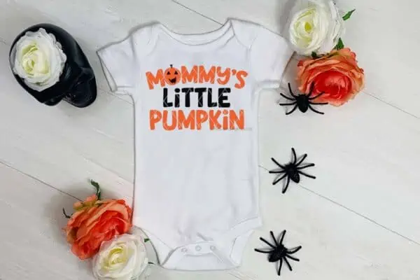Horizontal close up image of a white baby body suit with orange and black vinyl which reads mommy's little pumpkin with roses and spiders on wood background.