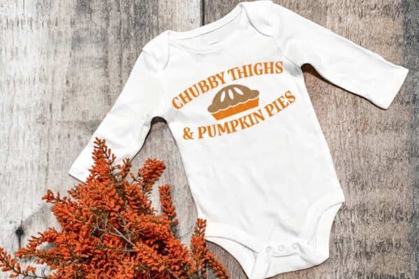 Horizontal image of white body suit on wood background which reads chubby thighs and pumpkin pies.