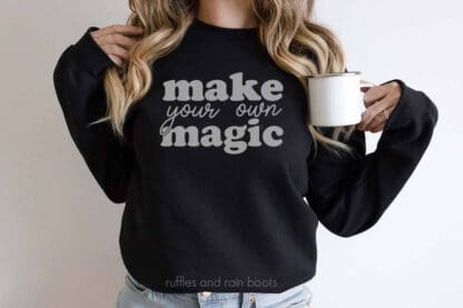 Horizontal image of woman holding cup wearing a black sweatshirt with make your own magic SVG in gray flock vinyl.