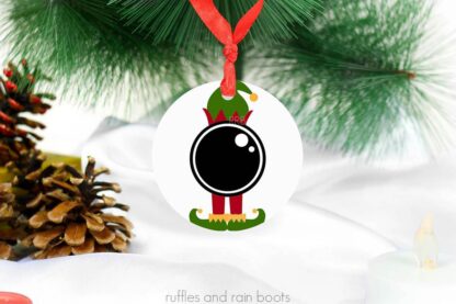 Close up white ceramic Christmas ornament with an elf cam made with Cricut and permanent vinyl on holiday background.