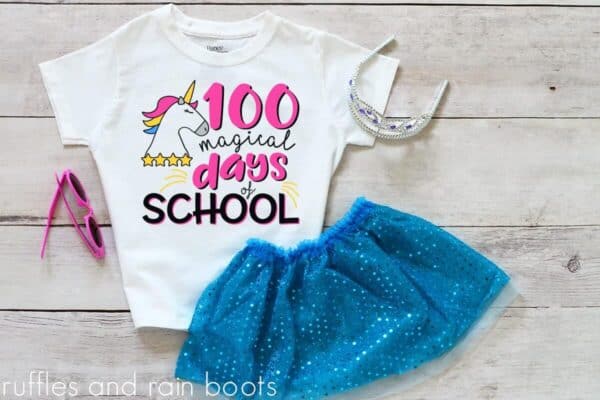 White wood background with blue tutu an white shirt which reads 100 magical days of school with a unicorn and stars.
