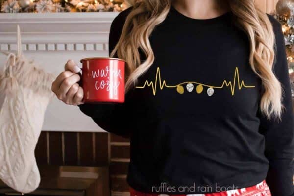 Horizontal image of woman holding a mug wearing a black t shirt with gold and silver foil Christmas lights heartbeat.