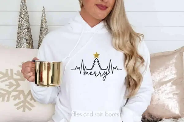 Horizontal image of woman holding gold mug wearing a white hoodie with black and gold vinyl tree heartbeat design.