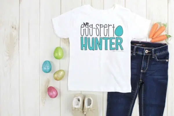 Horizontal image of a white shirt with blue and black eggspert hunter SVG with egg and bunny ears on wood background.