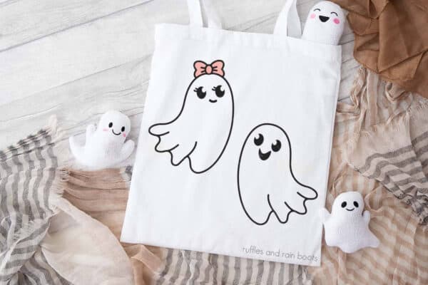 Horizontal image of a white trick or treat bag with ghost SVG outlines on a wood background with layered beige and tan scarves and tiny stuffed ghosts.