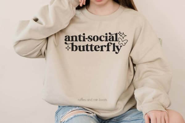 Horizontal image of woman in tan sweatshirt made with Antisocial Butterfly SVG and black heat transfer vinyl on Cricut.