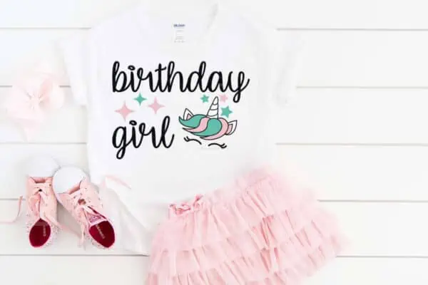 Horizontal close up of pink, ruffled skirt, pink shoes, and white t shirt on wood which reads birthday girl and a unicorn face SVG with stars.