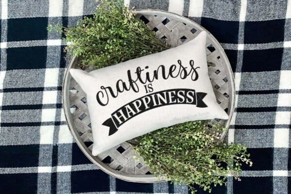 Horizontal image of plaid blanket with white basket and greens holding white rectangle pillow which reads craftiness is happiness in vinyl.