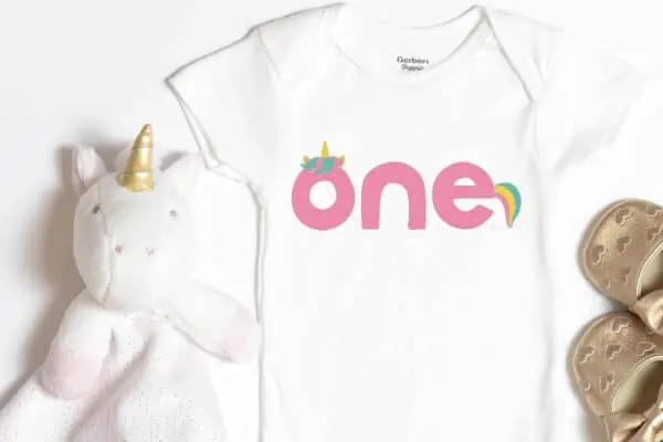 Horizontal image of white baby bodysuit with unicorn SVG spelling out one in pink, yellow, and teal.