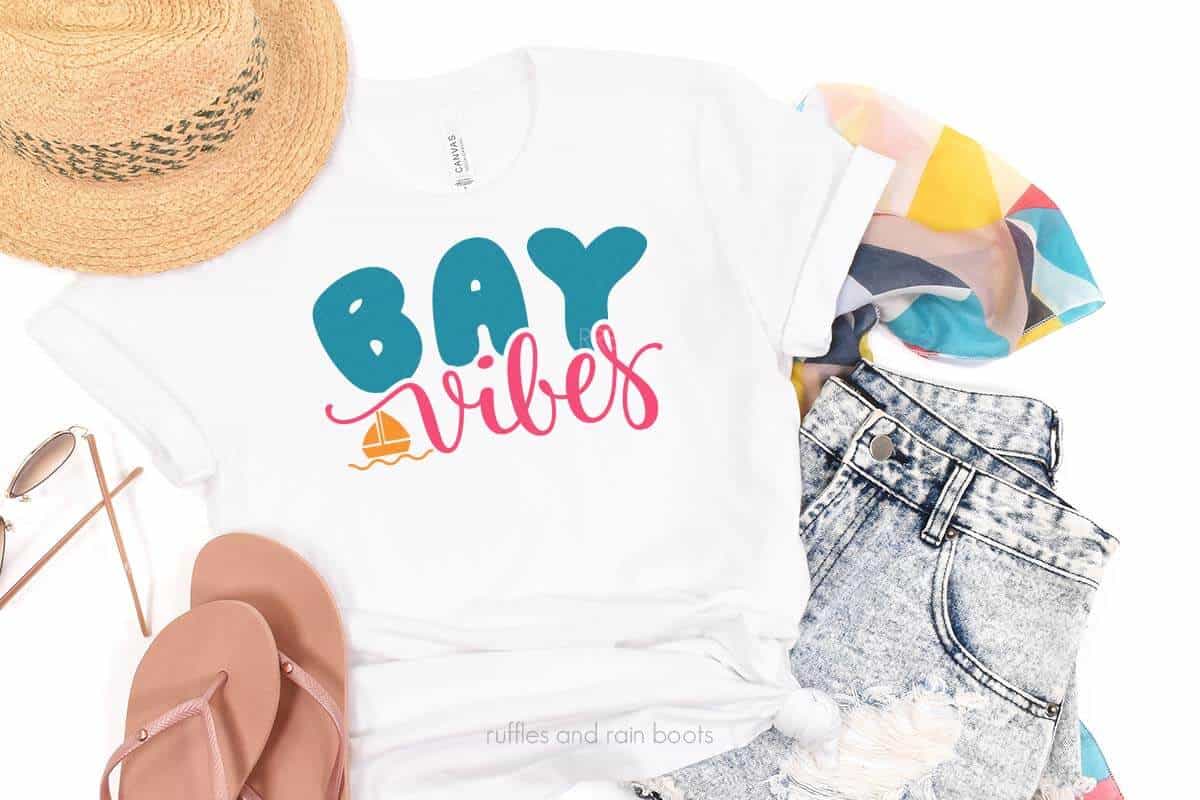Horizontal image of white t-shirt which reads bay vibes on a white background with hat, shorts, sandals, and sunglasses.