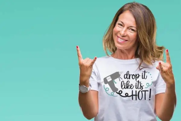 Horizontal image of smiling woman standing in front of teal wall wearing a white t-shirt which reads drop it like it's hot with a glue gun.