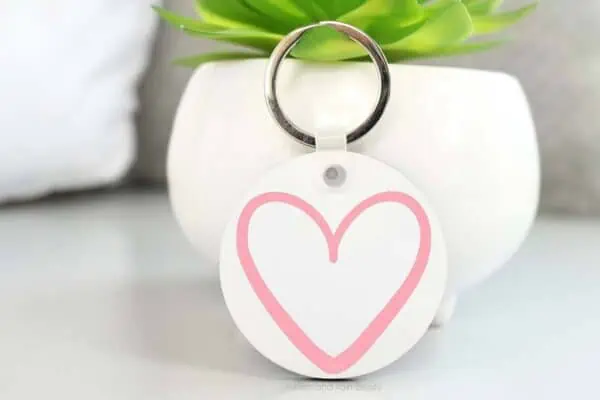 Horizontal image of a white acrylic keychain with a pink hand drawn heart in front of small plant.
