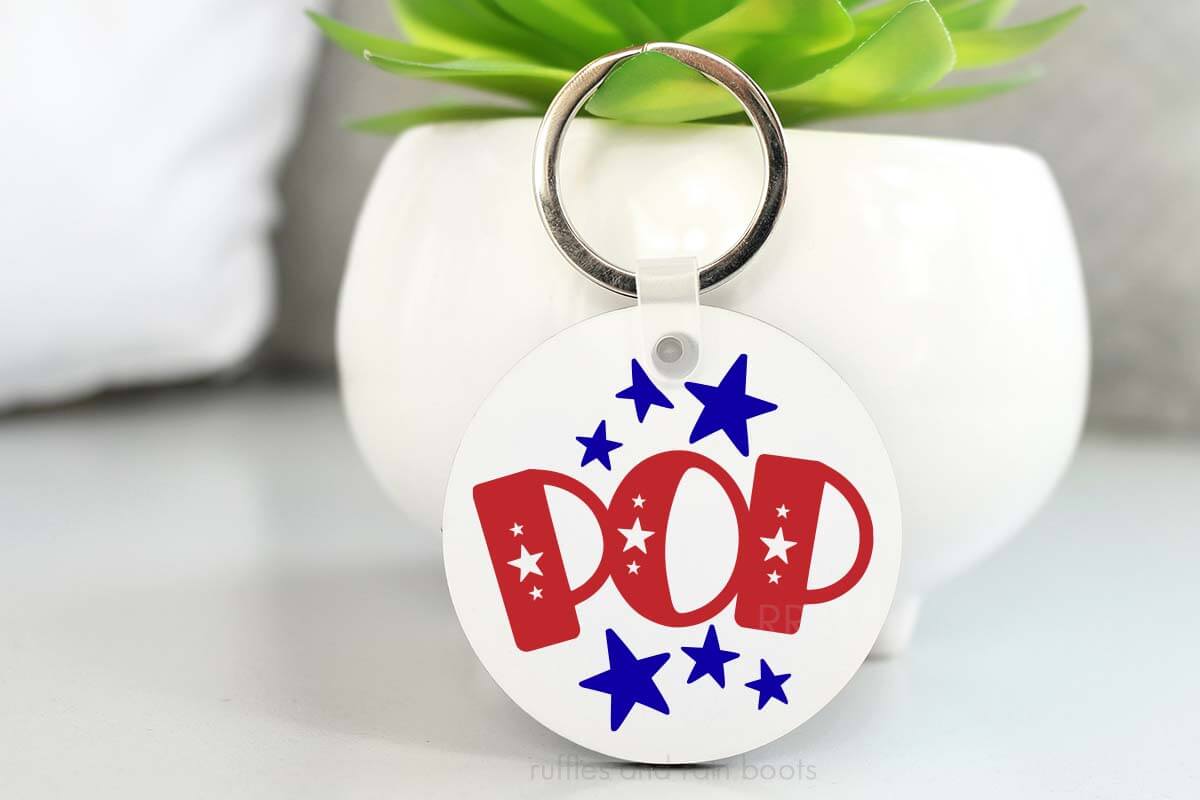 Horizontal image of white plant propping up white keychain with POP and Stars SVG for Independence Day.