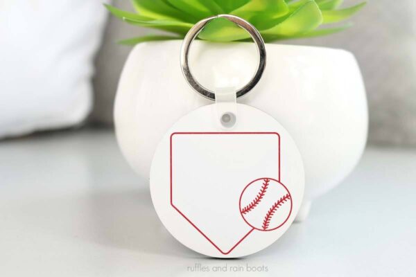 Horizontal image of white acrylic keychain with baseball SVG and home plate cut file leaning against a small plant.