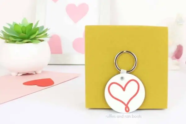 Horizontal image of a white keychain with a hand drawn heart SVG free cut file leaned against a yellow box.