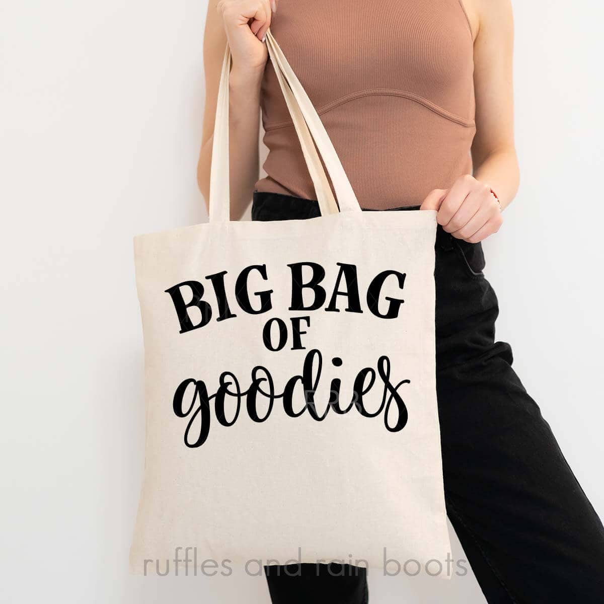 Vertical image of woman in black jeans and brown top holding canvas tote which reads big bag of goodies.