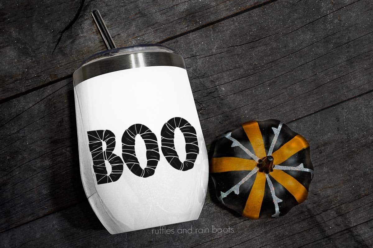 Horizontal image of white tumbler which reads boo in mummy style on black wood with painted pumpkin.