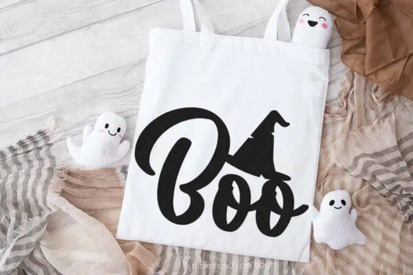 Horizontal image of a white tote bag on neutral Halloween background which reads boo with a witch hat in vinyl.